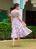 Woman in a floral dress standing outdoors with greenery and a building in the background Glanzavi