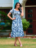 Woman wearing a blue floral dress standing outdoors with greenery and a building in the background Glanzavi