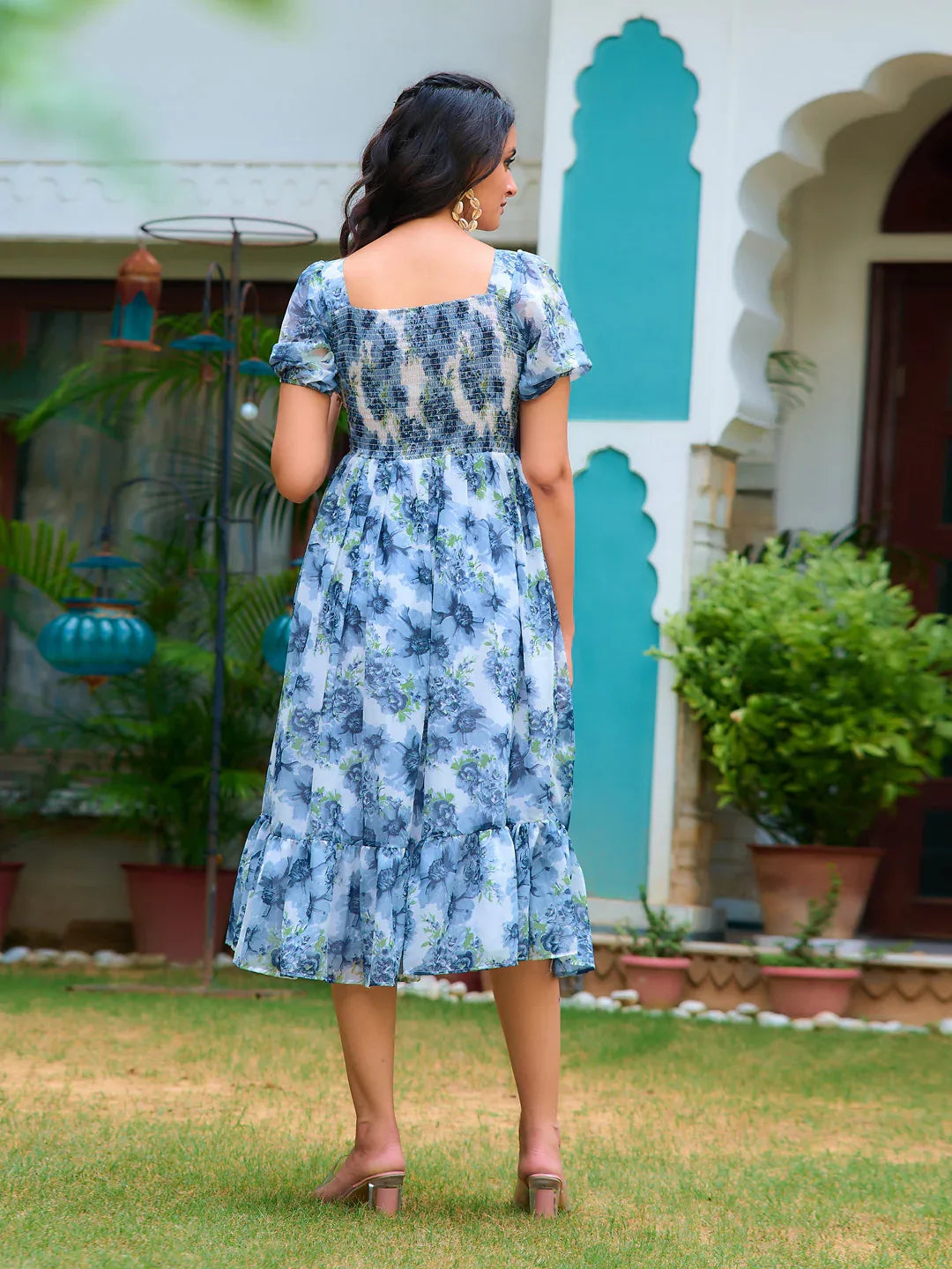 Woman in a blue floral dress standing in front of a building with greenery Glanzavi