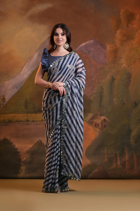 A woman modeling a grey striped saree with mirror and sequin embellishments, standing against a traditional painted backdrop Glanzavi