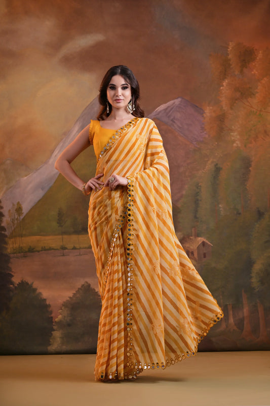 A woman modeling a yellow striped saree with mirror and sequin embellishments, standing against a traditional painted backdrop Glanzavi