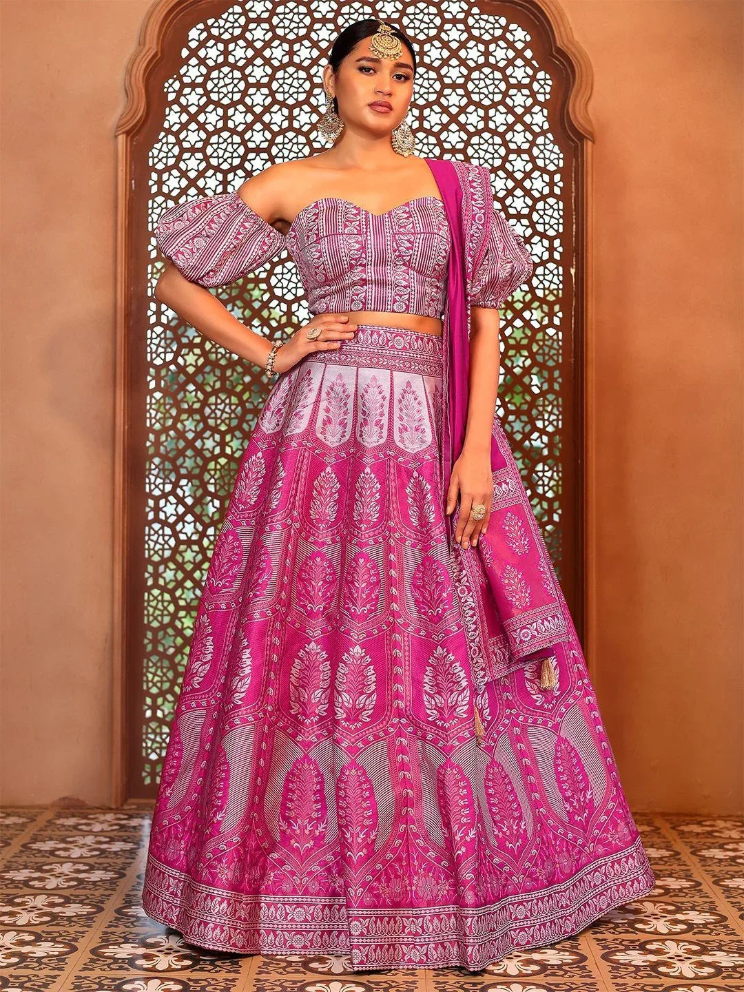 Woman wearing a pink and purple traditional outfit with intricate patterns against a decorative wall Glanzavi.