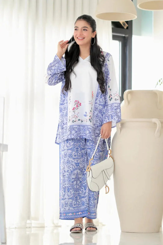 Woman in a blue floral outfit standing in a bright room with white curtains and a vase Glanzavi