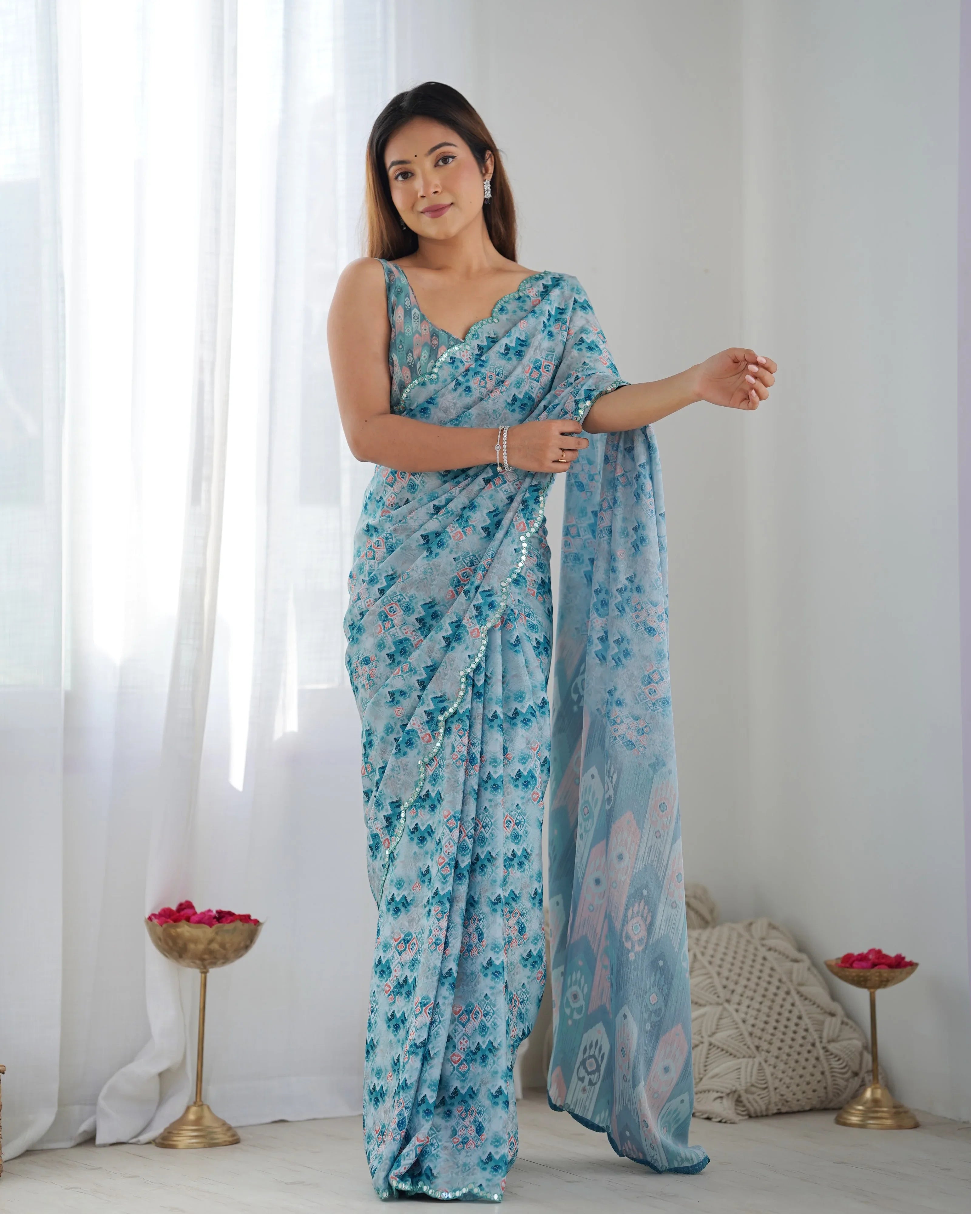 A woman modeling a sky blue ethnic printed georgette saree with scalloped lace and a blouse piece, standing against a backdrop with curtains and decorative items Glanzavi