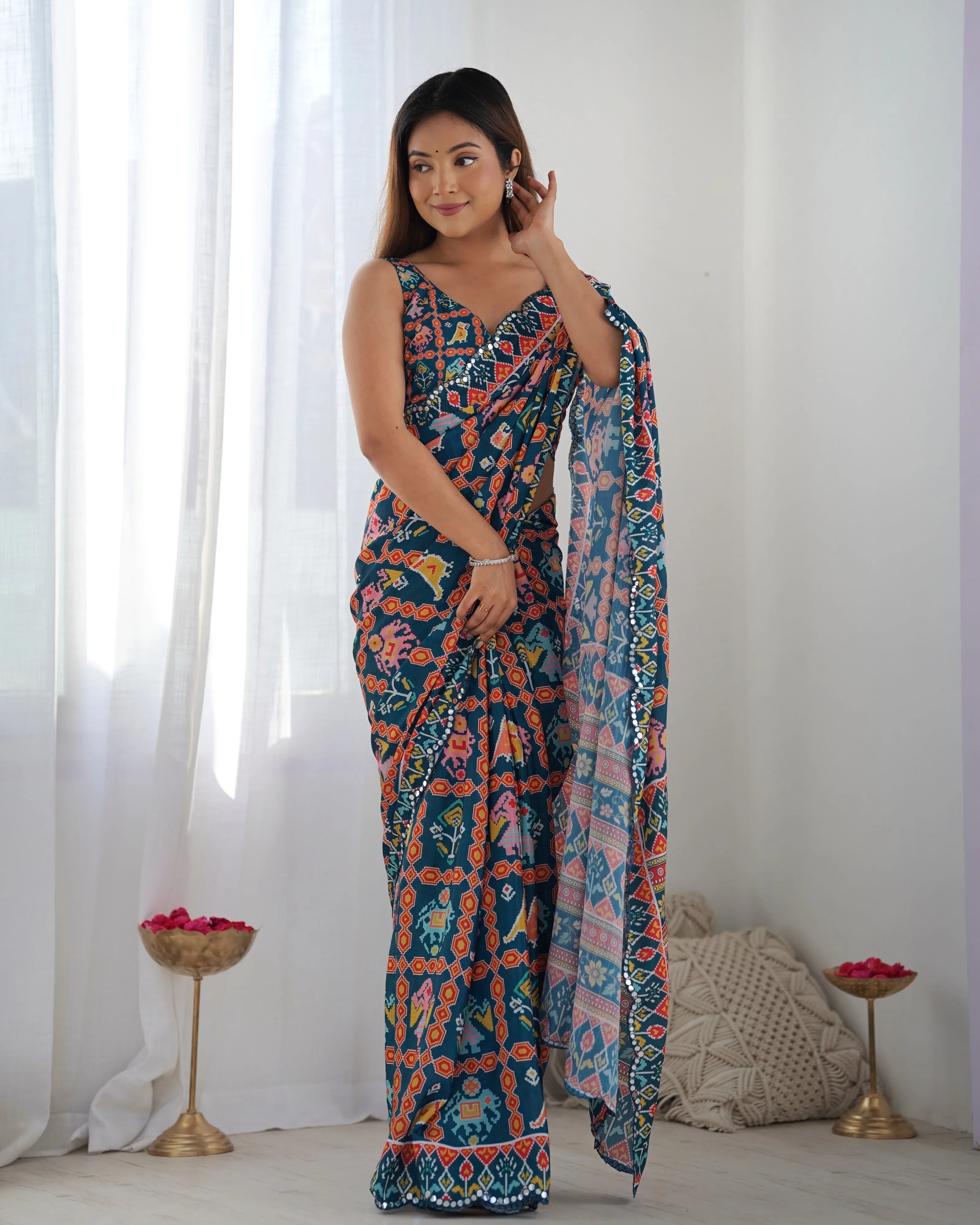 Woman wearing a colorful patterned saree in a room with white curtains and decorative items Glanzavi
