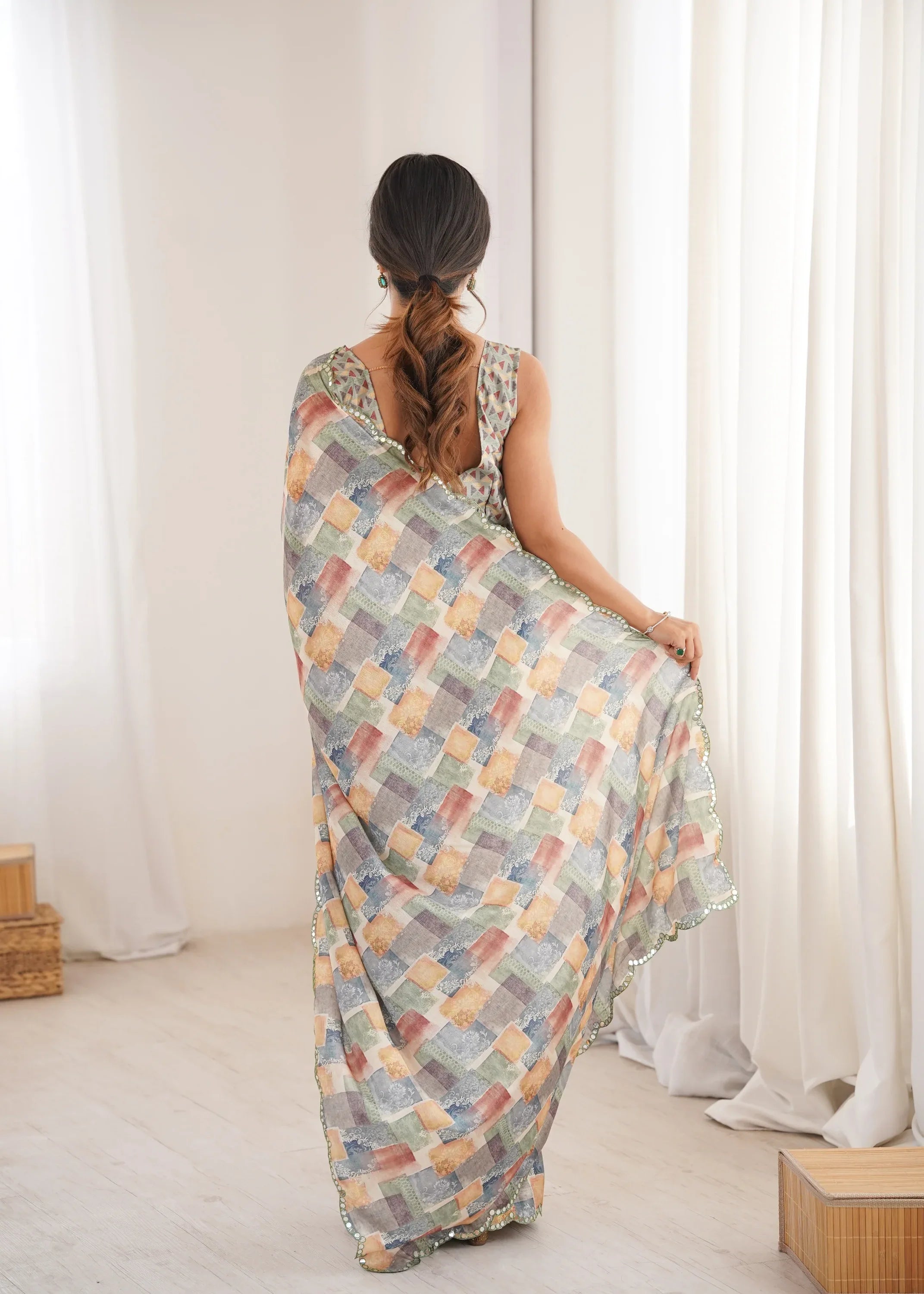 Woman wearing a colorful patterned saree sitting in a room with white curtains Glanzavi