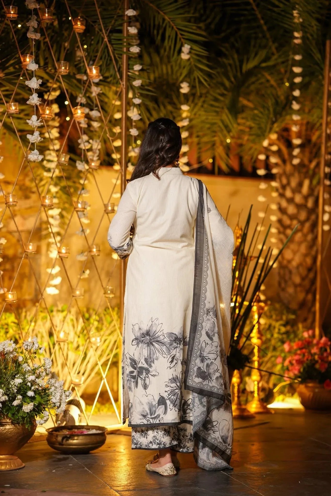 Woman in a white saree with black patterns standing in front of decorative lights and plants Glanzavi