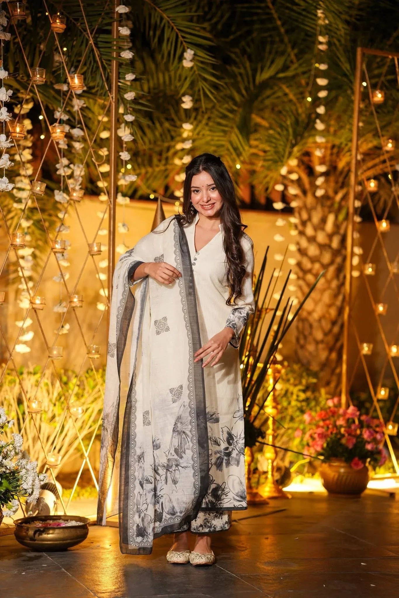 Woman in a white traditional outfit with a floral pattern, standing in a decorated outdoor setting with lights and plants Glanzavi