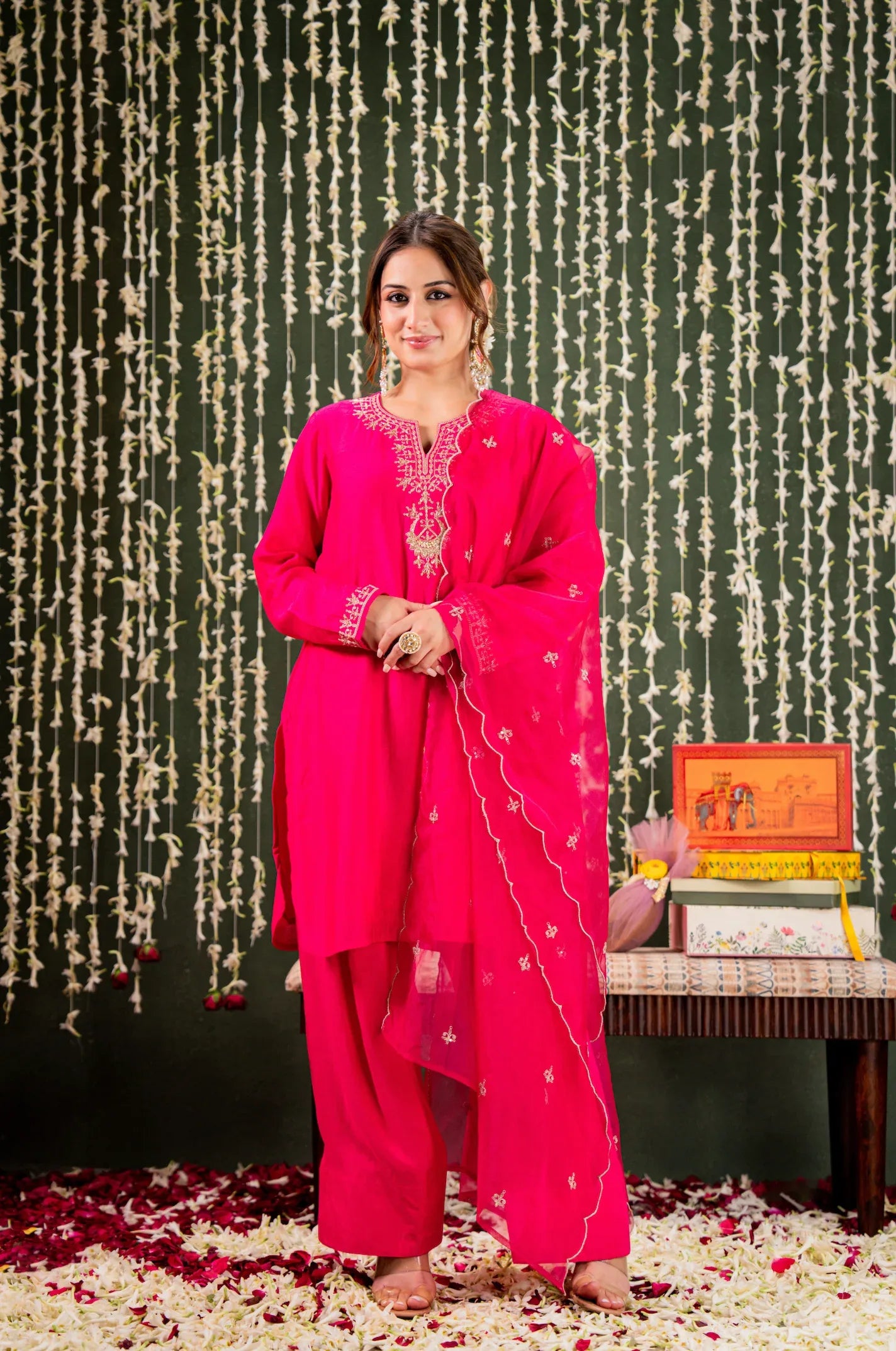Woman in a bright pink traditional outfit standing in front of a decorative backdrop with floral decorations on the floor Glanzavi