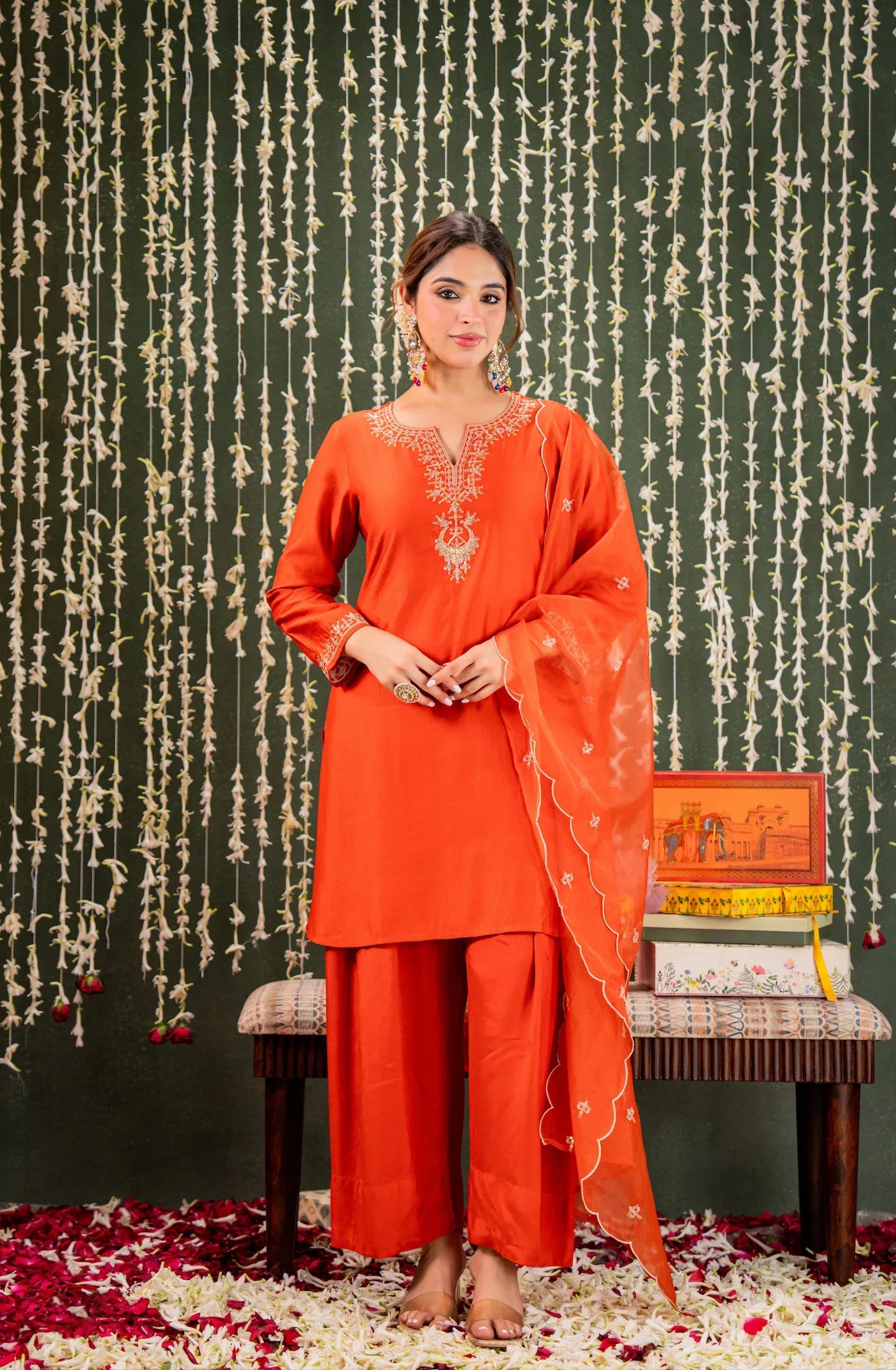 Woman in an orange traditional outfit standing against a decorative background with floral arrangements Glanzavi