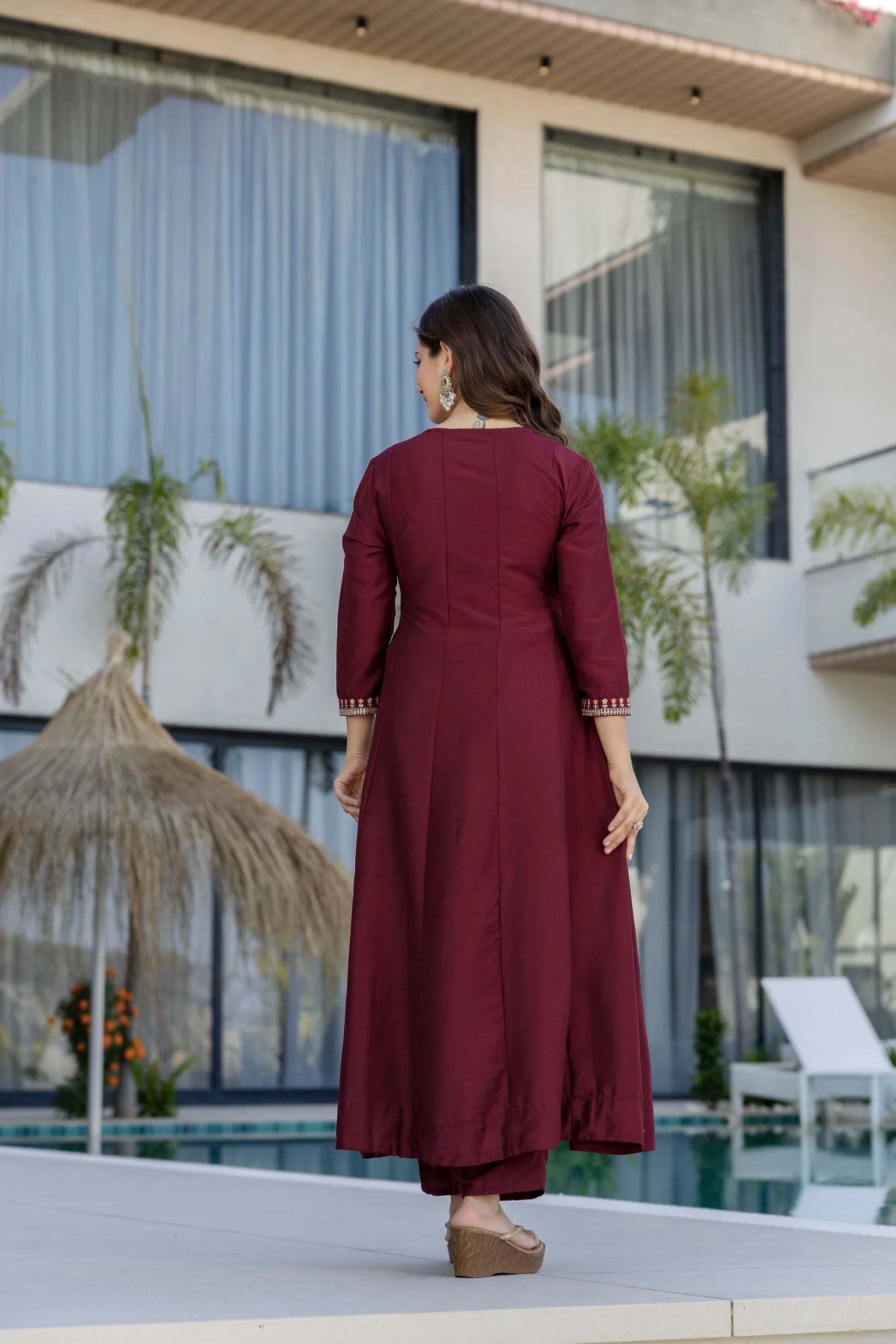 Woman in a maroon outfit standing by a poolside with a modern building and plants in the background Glanzavi