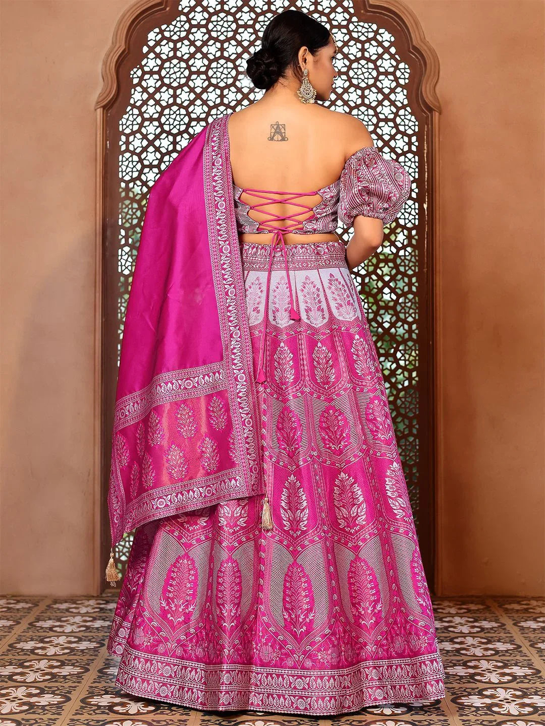 Woman wearing a pink and gray embroidered traditional outfit with a matching dupatta, standing in front of an ornate wooden door Glanzavi.