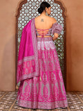 Woman wearing a pink and gray embroidered traditional outfit with a matching dupatta, standing in front of an ornate wooden door Glanzavi.