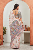 Woman in a traditional saree with a colorful pattern, standing in a room with a white wall and wooden shelf Glanzavi