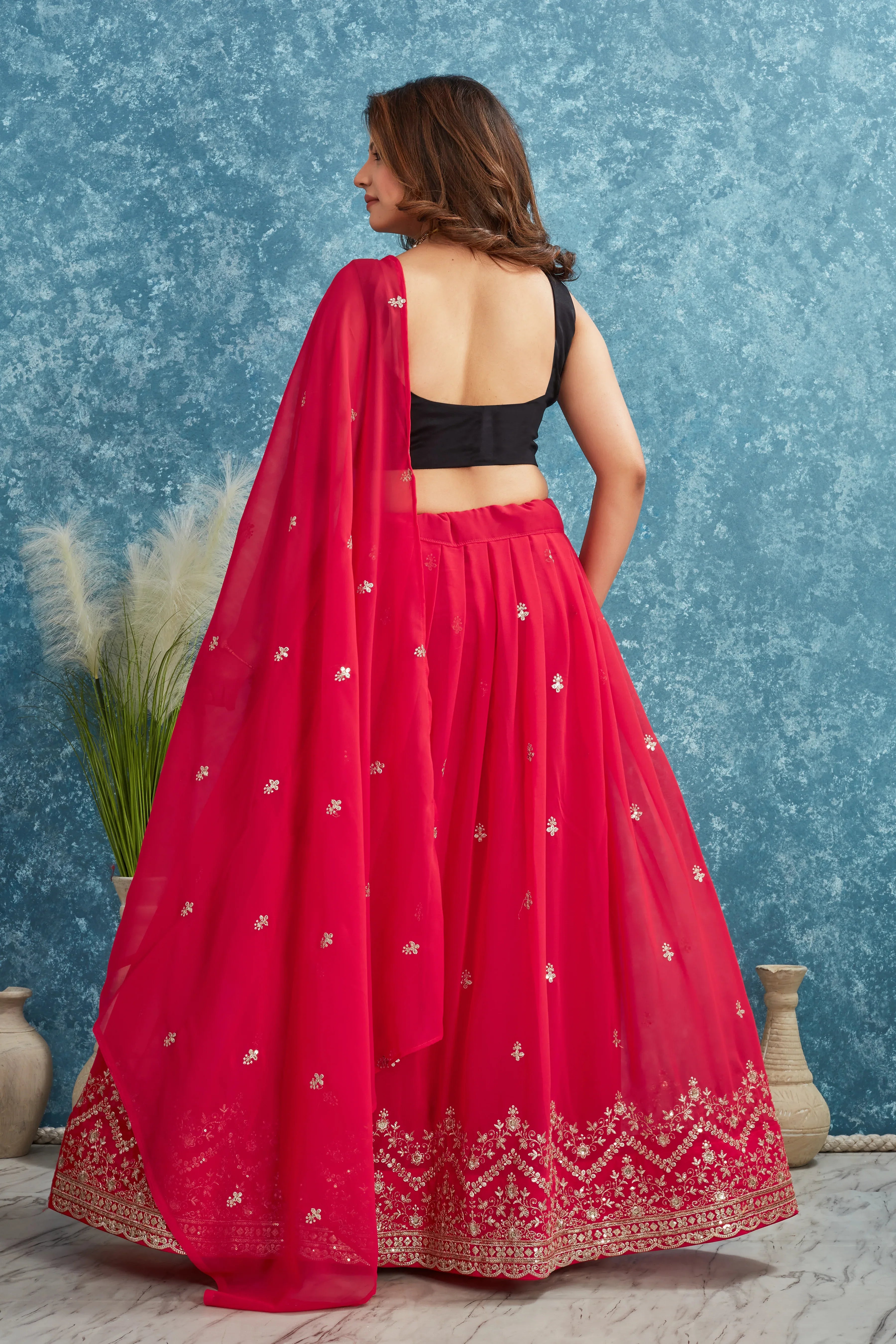 Woman wearing a red lehenga with a black blouse against a blue textured wall Glanzavi