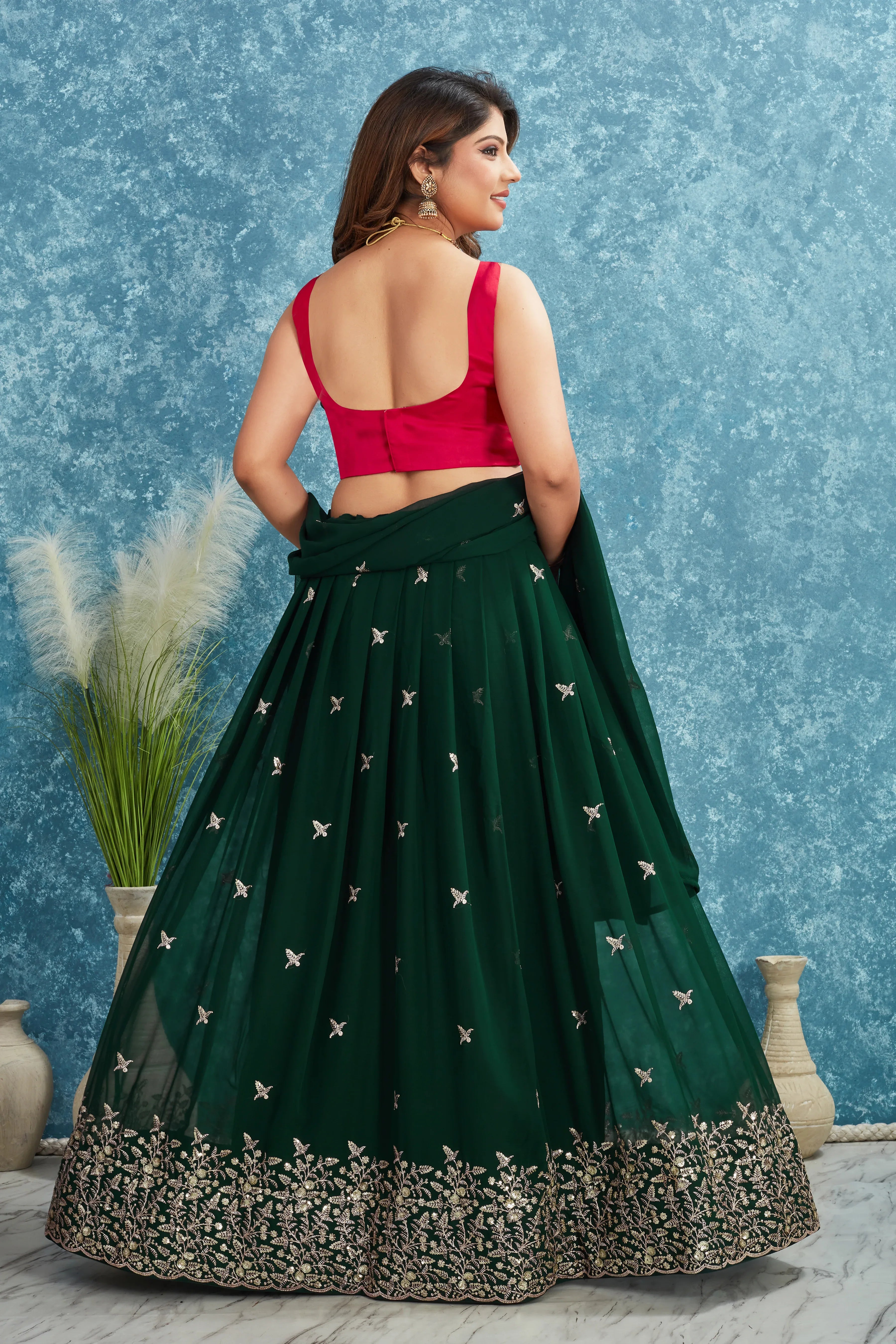 Woman wearing a green lehenga with a red blouse against a blue textured wall Glanzavi