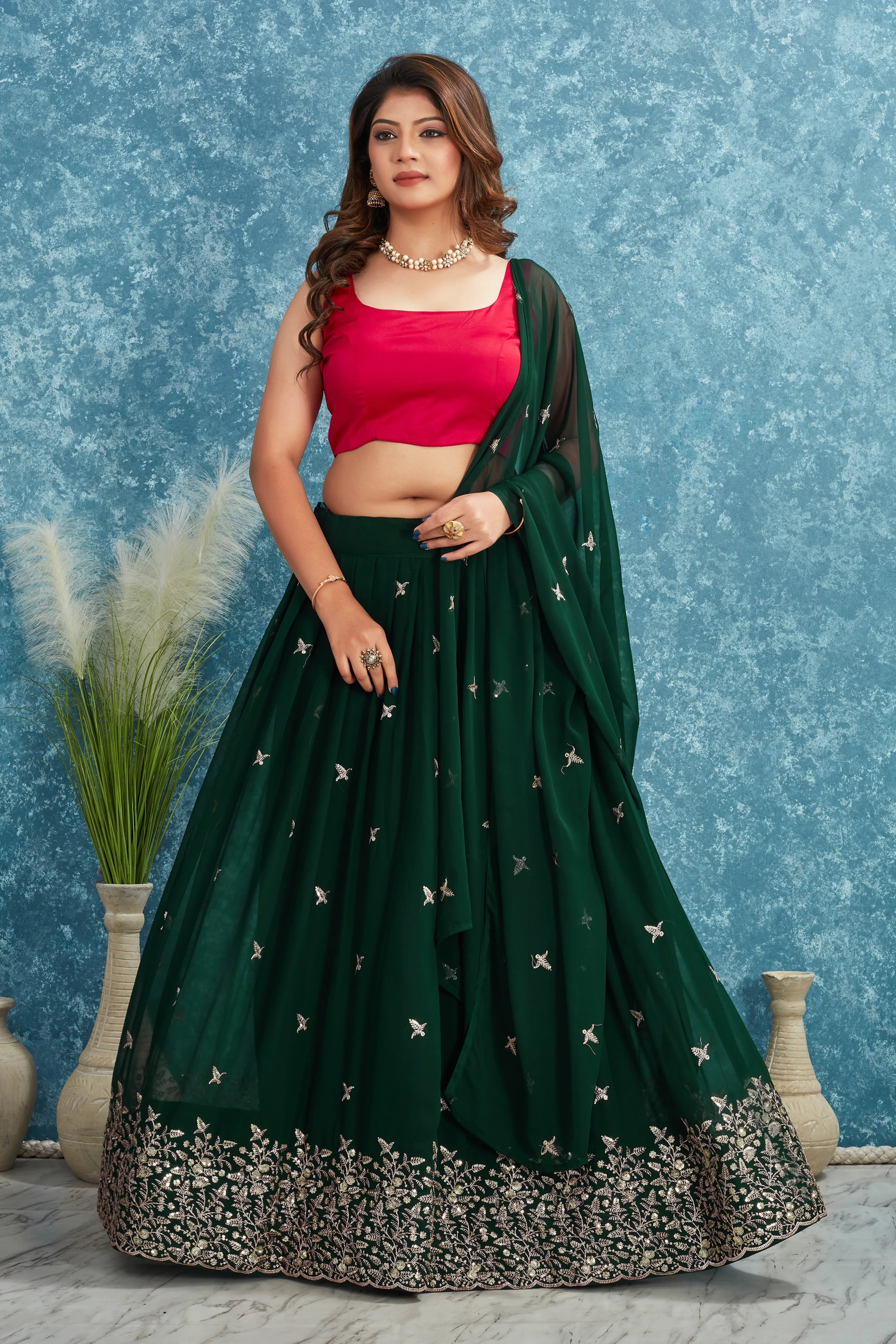 Woman wearing a green lehenga with pink blouse against a blue textured wall Glanzavi
