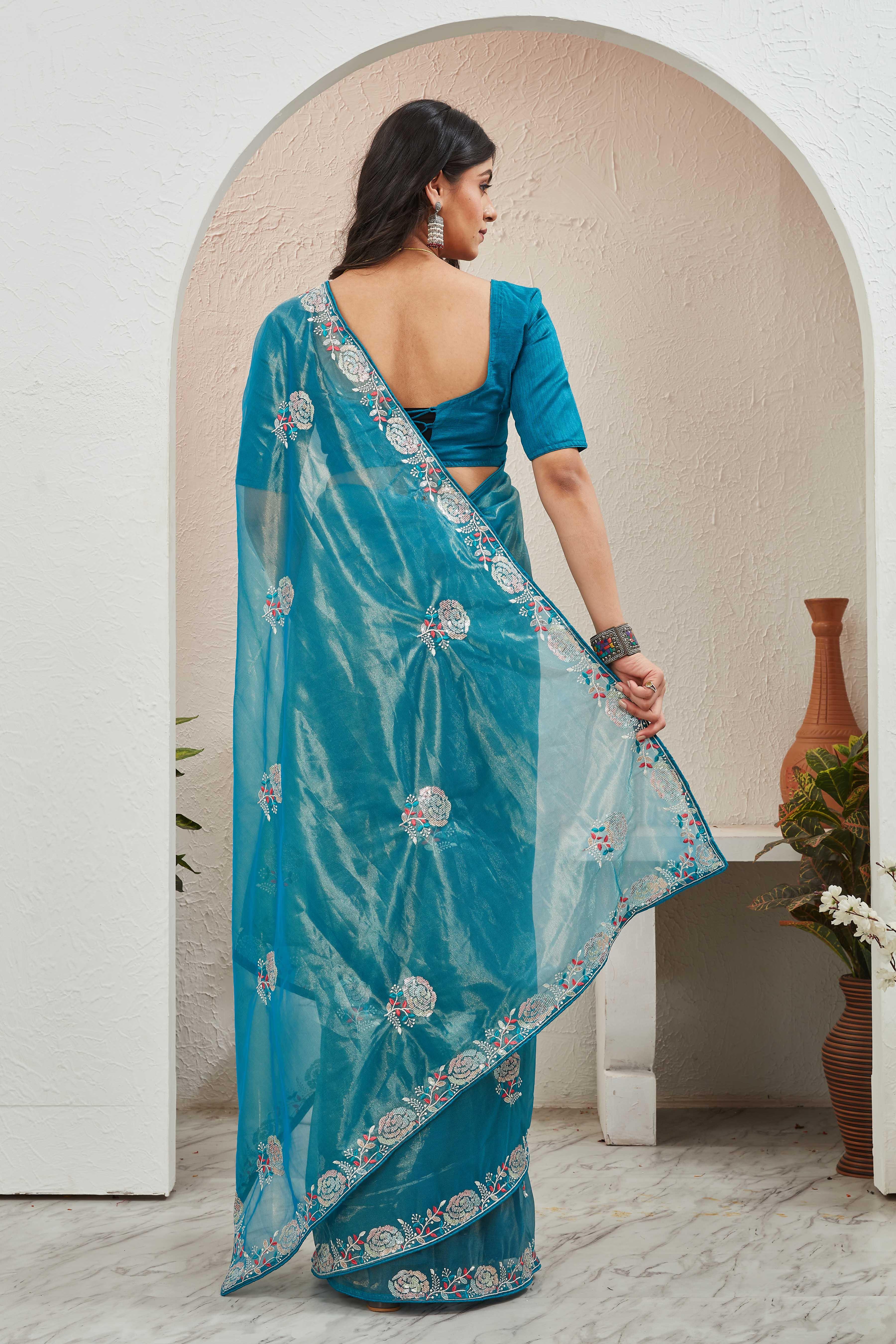 Woman wearing a blue saree with a floral pattern, standing in a room with a white wall and a vase Glanzavi.