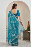 Woman wearing a blue saree with a floral pattern, standing in a room with a white wall and a vase Glanzavi.