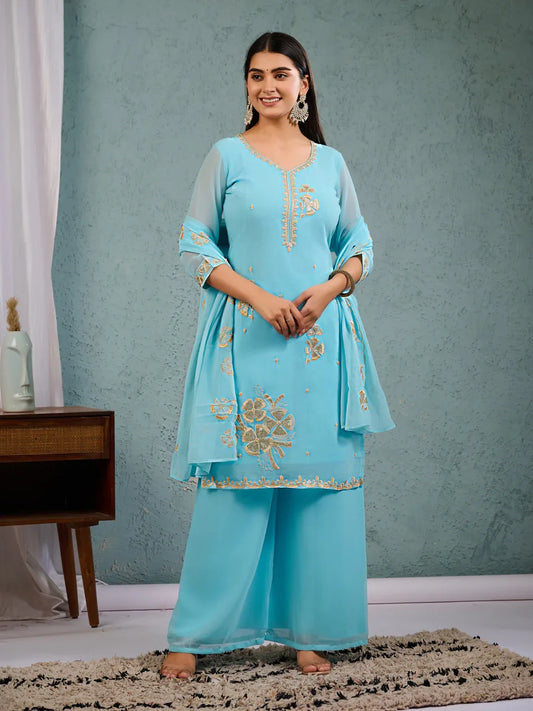 Woman wearing a light blue embroidered traditional outfit against a gray wall Glanzavi