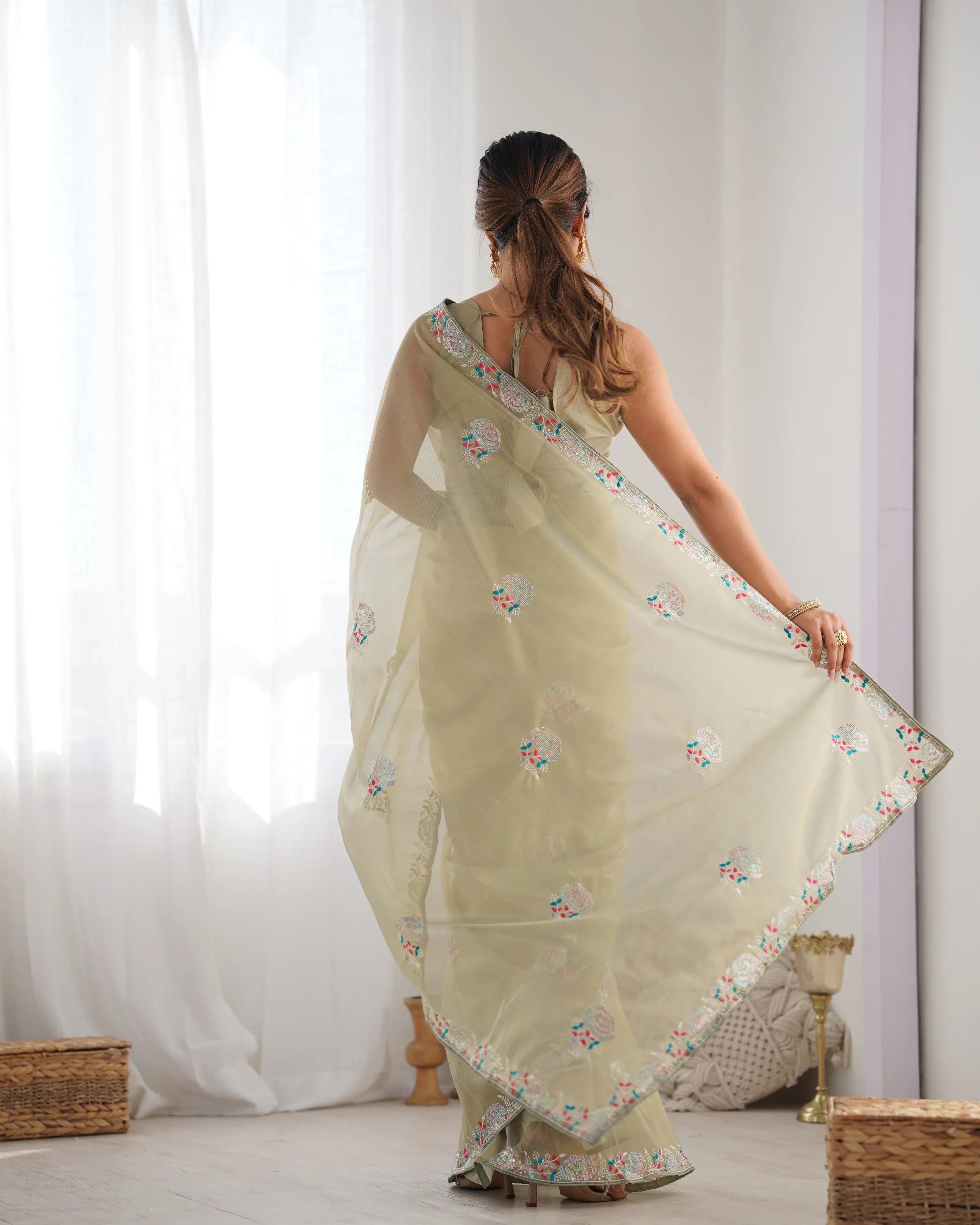 Woman wearing a light green saree with floral patterns in a room with white curtains Glanzavi
