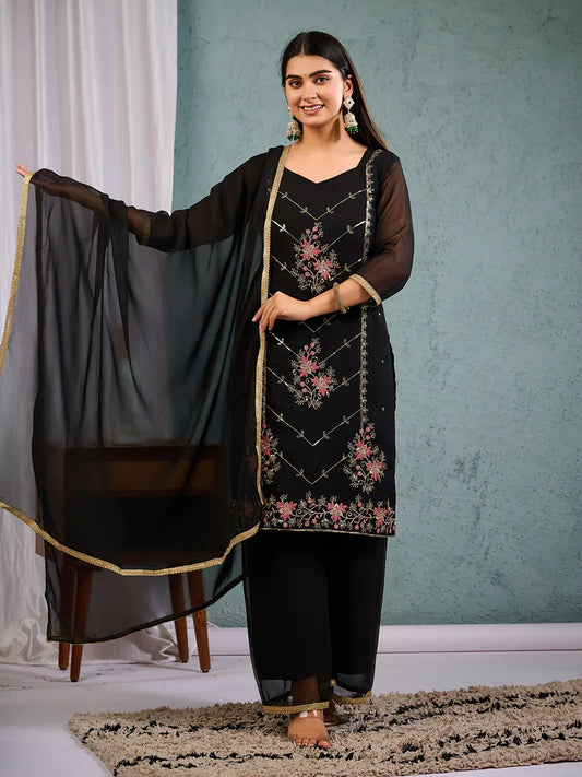 A woman standing in a room with a light blue background, wearing an elegant black georgette suit with intricate thread embroidery and a sheer dupatta with a golden border Glanzavi