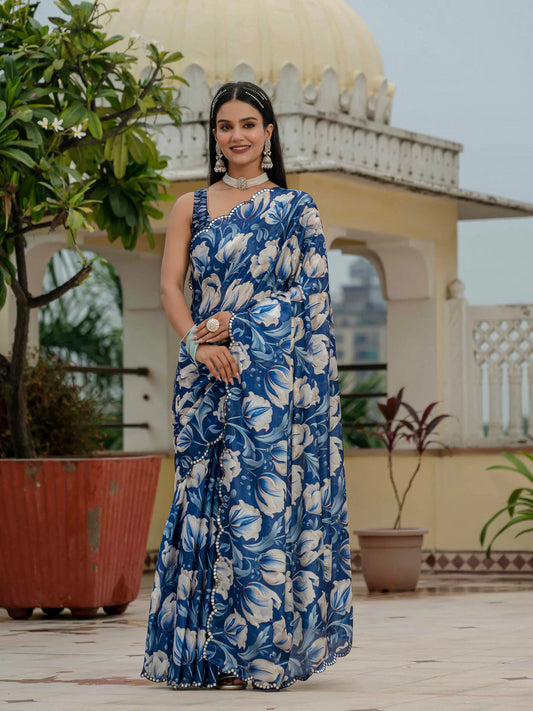 Woman in a blue floral saree standing outdoors with architectural and plant elements in the background Glanzavi