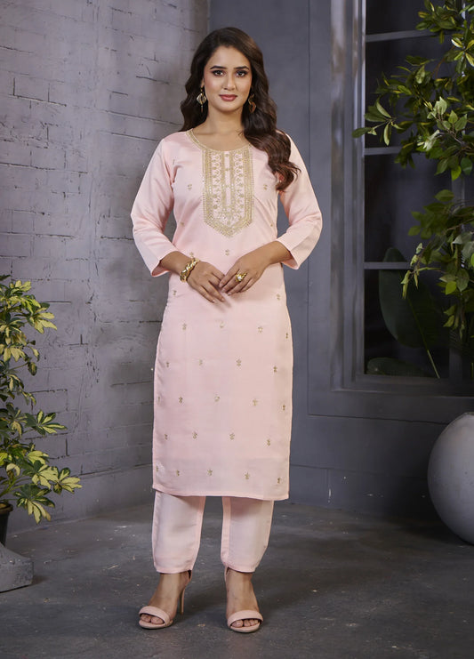 Woman wearing a light pink traditional outfit with embroidery, standing against a gray brick wall Glanzavi