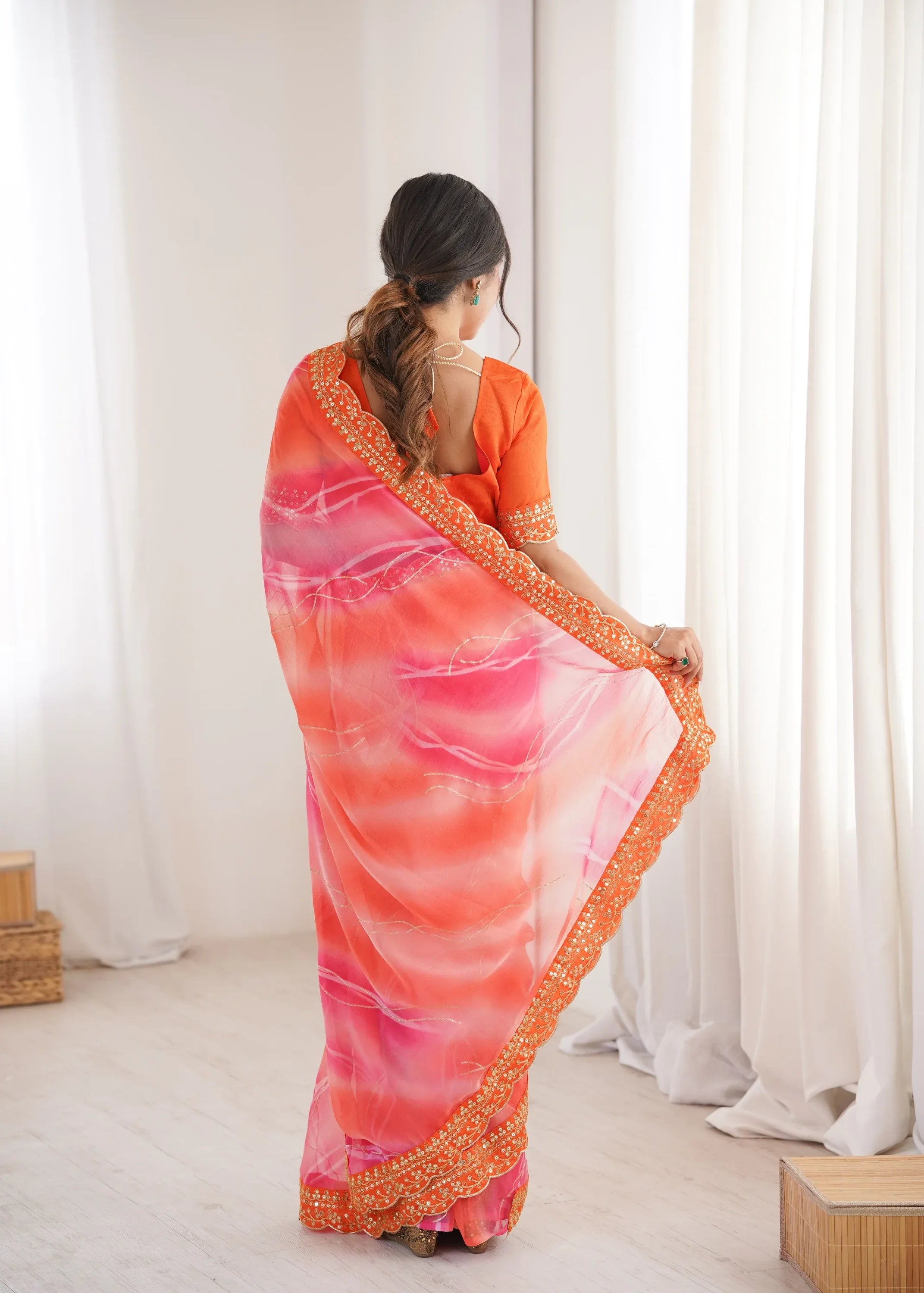Woman wearing a vibrant saree with a pink and orange gradient, standing against a white curtain background Glanzavi.