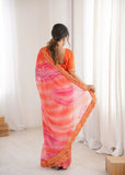 Woman wearing a vibrant saree with a pink and orange gradient, standing against a white curtain background Glanzavi.