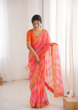 Woman wearing a vibrant orange and pink saree with gold border indoors Glanzavi.