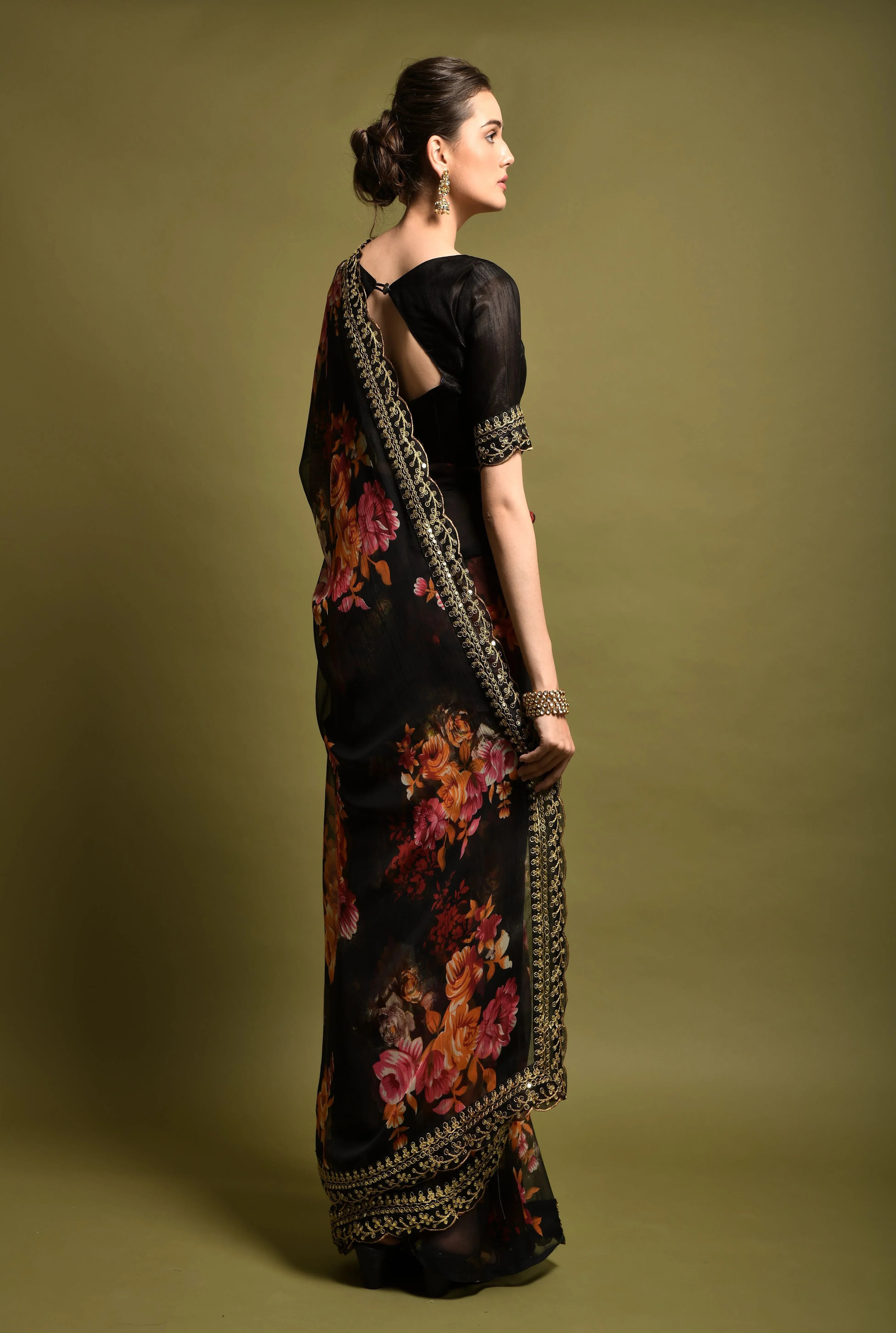 Woman wearing a black floral saree with gold border against a green background Glanzavi
