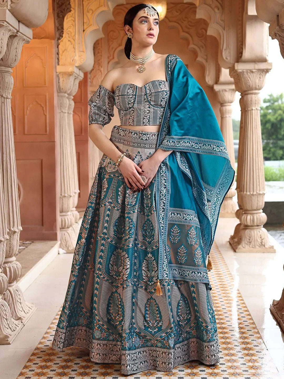 Woman in a traditional blue and gray embroidered outfit standing in an ornate architectural setting Glanzavi.