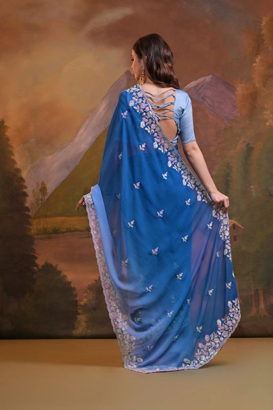 Woman wearing a blue saree with floral patterns in front of a scenic background Glanzavi
