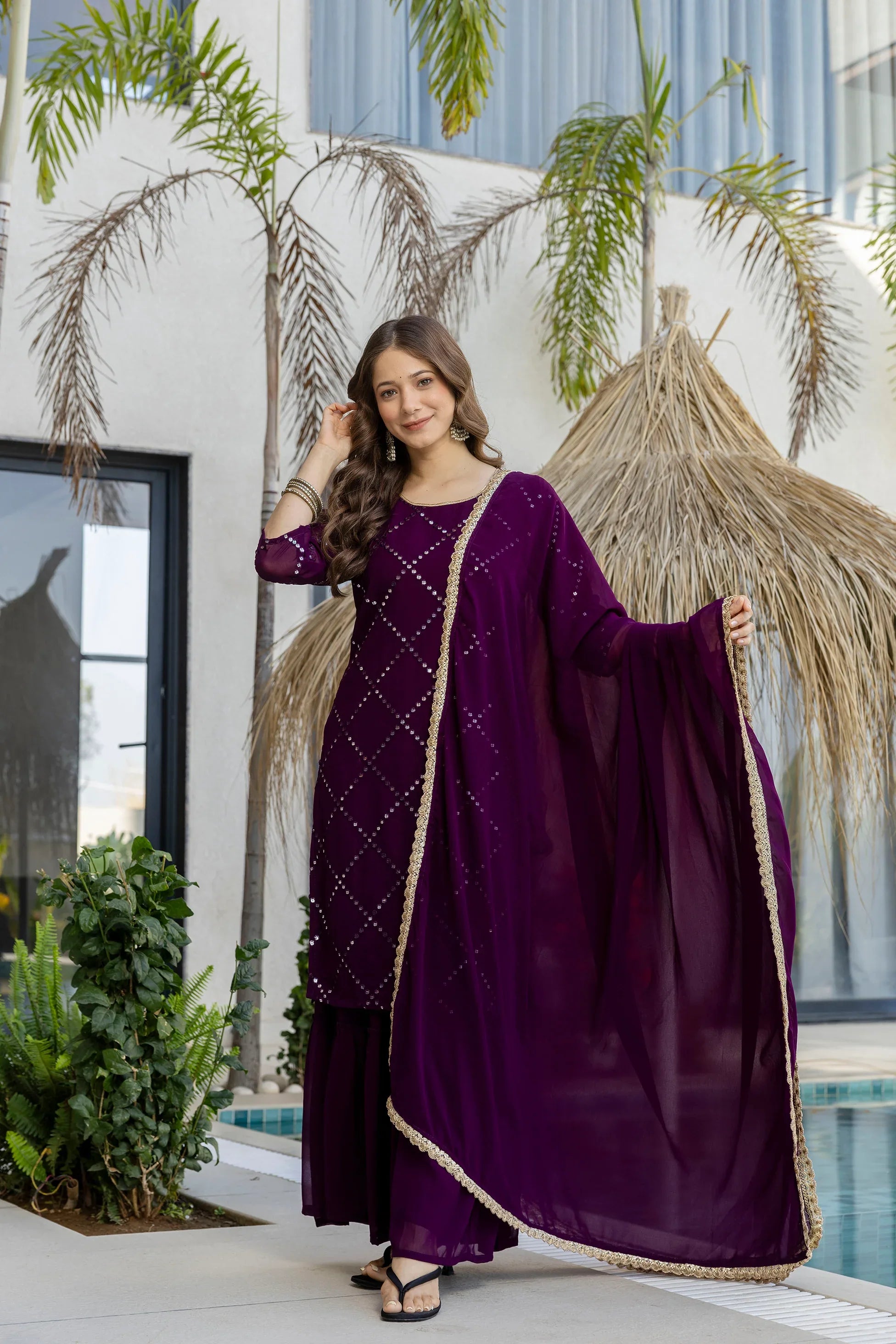 Woman in a purple traditional outfit standing outdoors with plants and a building in the background Glanzavi