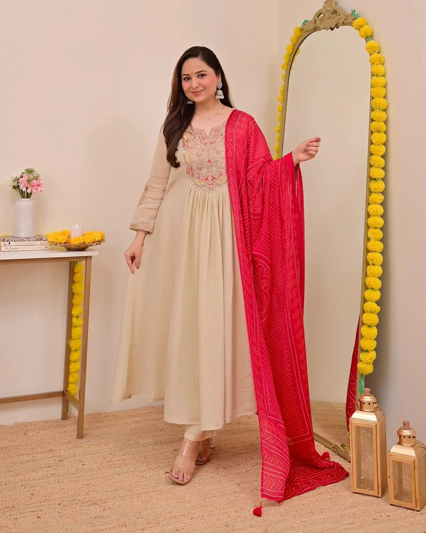 Woman in a beige anarkali suit with a red dupatta standing next to a decorative mirror Glanzavi