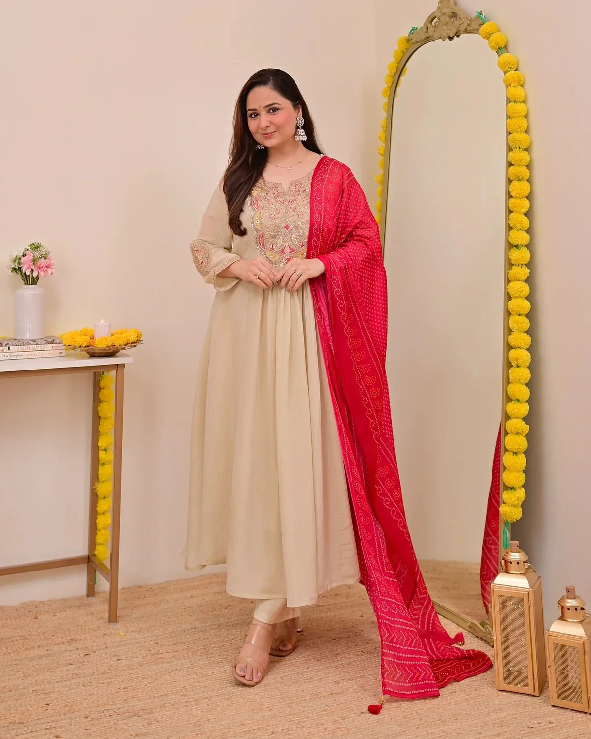 Woman in a beige outfit with a red dupatta standing in front of a decorated mirror Glanzavi