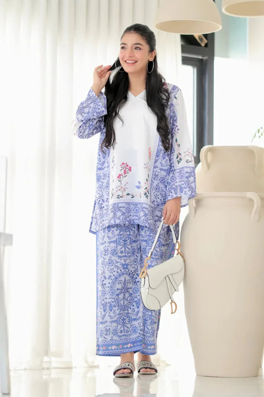 Woman in a blue floral outfit standing in a bright room with white curtains and a vase Glanzavi