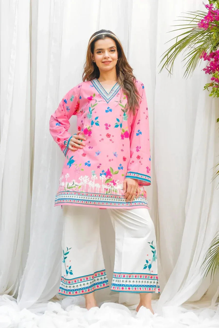 Woman wearing a pink embroidered shirt and white pants against a white curtain background Glanzavi