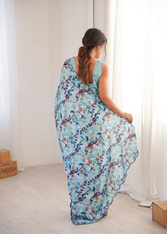 Woman wearing a blue floral saree in a bright room with white curtains Glanzavi.