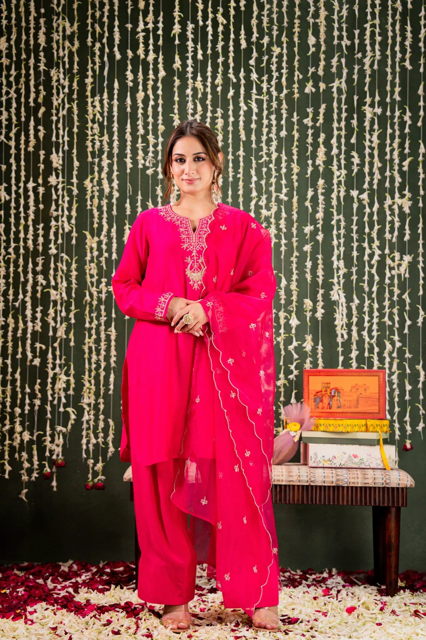 Woman in a bright pink traditional outfit standing next to a small table with decorative items against a dark background Glanzavi