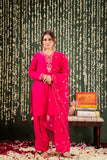Woman in a bright pink traditional outfit standing next to a small table with decorative items against a dark background Glanzavi
