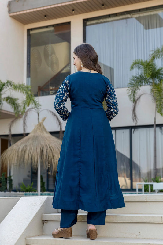 Woman in a blue dress standing on steps with a building and plants in the background Glanzavi
