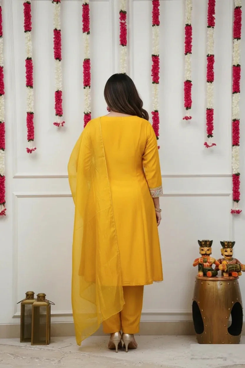 Woman in a yellow traditional outfit standing in front of a decorative wall with floral arrangements Glanzavi